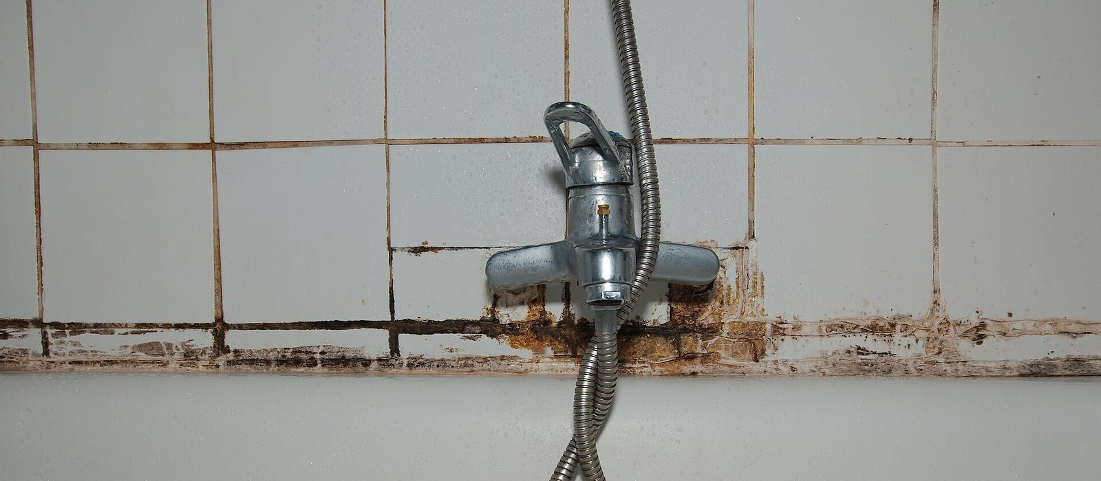 Versleten douchekraan met verkleurde voegen op witte tegels in een badkamer die gerenoveerd wordt